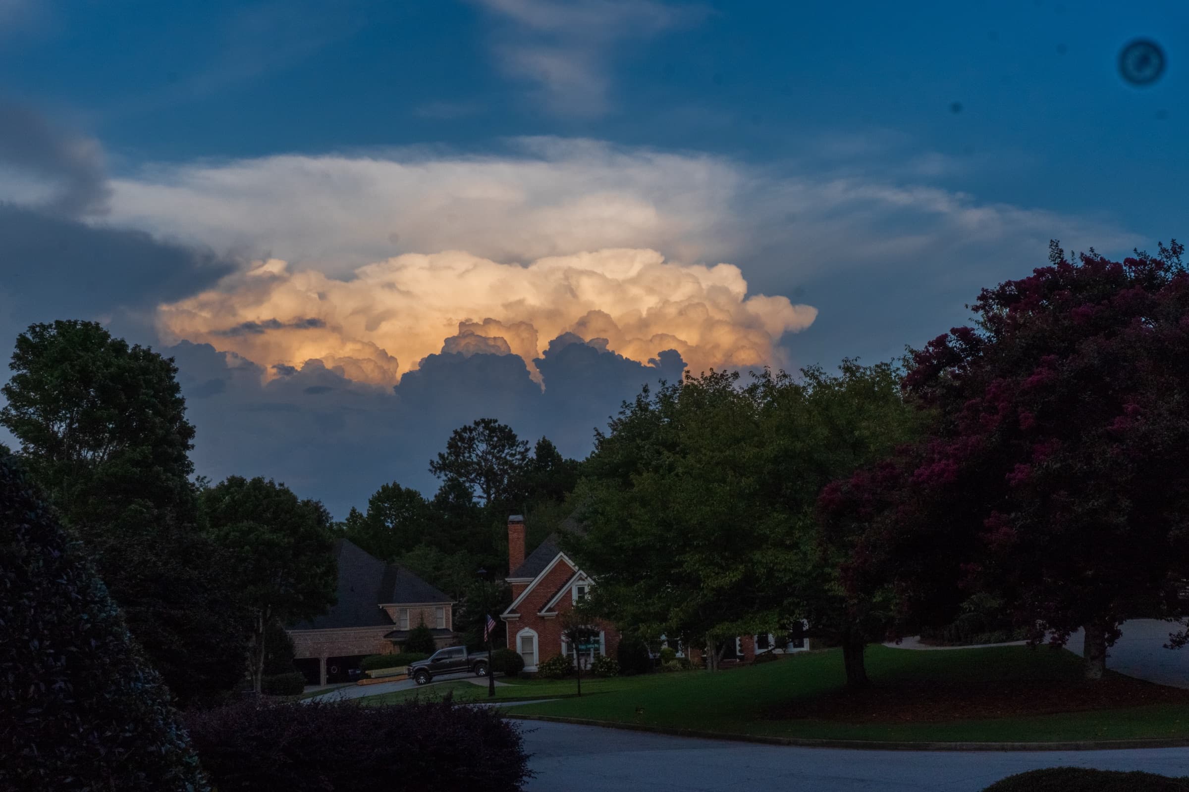 A towering cumulonimbus cloud lit gold by the setting sun above a suburban street lined with trees